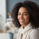 A happy young woman with dark, curly hair smiles genuinely in a soft-focus portrait headshot, conveying satisfaction.